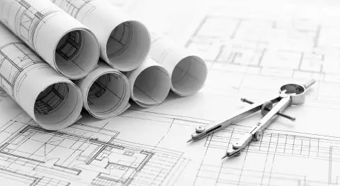 A black and white photograph showcasing a stack of rolled-up architectural blueprints placed on the upper-left side. The background features open, detailed construction floor plans spread flat across a surface, with a metallic drafting compass tool resting on top of them in the foreground.