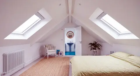 A brightly lit, modern loft conversion bedroom with white walls and a vaulted ceiling featuring exposed central beams. Symmetrical skylight windows on either side of the sloped roof fill the space with natural light. The room is furnished with a bed made with light-colored bedding, a woven floor rug, a white armchair, and a potted plant. An open doorway at the far end reveals an en-suite bathroom with a blue pedestal sink and a round mirror.